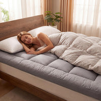 Woman lying on a bed with a gray mattress topper and beige comforter in a bright room.